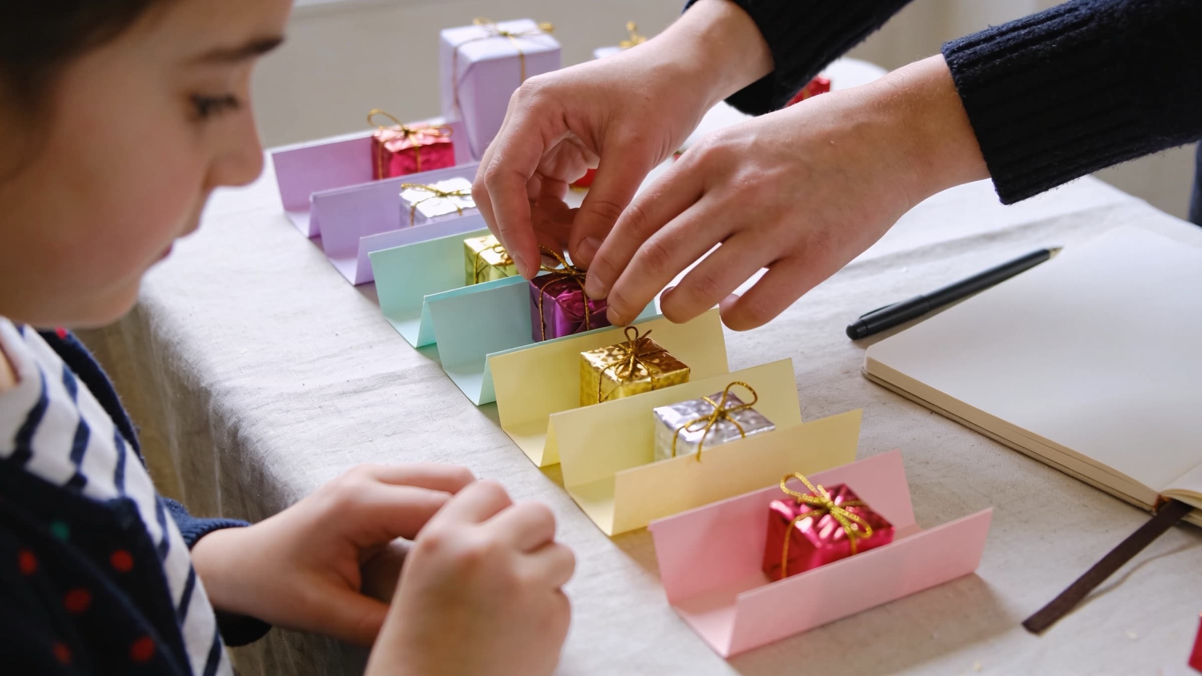 Des mains d’adulte rangent de petits cadeaux emballés de papier métallisé dans des supports en papier pastel pliés, sous le regard d’un enfant en marinière